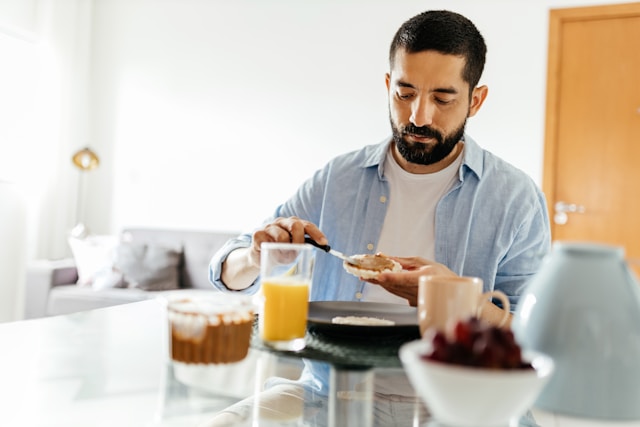 Man in a blue shirt spreading jam on toast at a breakfast table with a glass of orange juice and a bowl of grapes, focused and serene in a well-lit room.