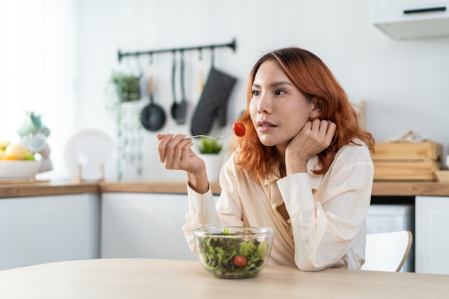 Woman in a beige blouse eating a cherry tomato from a bowl of salad in a modern kitchen.