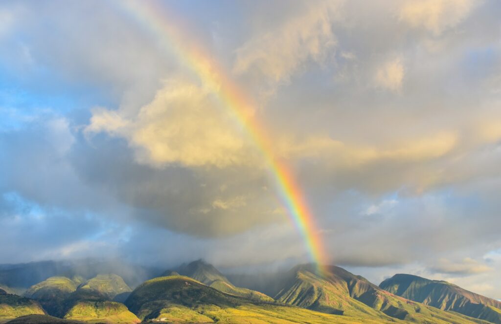 Vibrant rainbow over green mountains in Hawaii, with dramatic clouds in the sky.