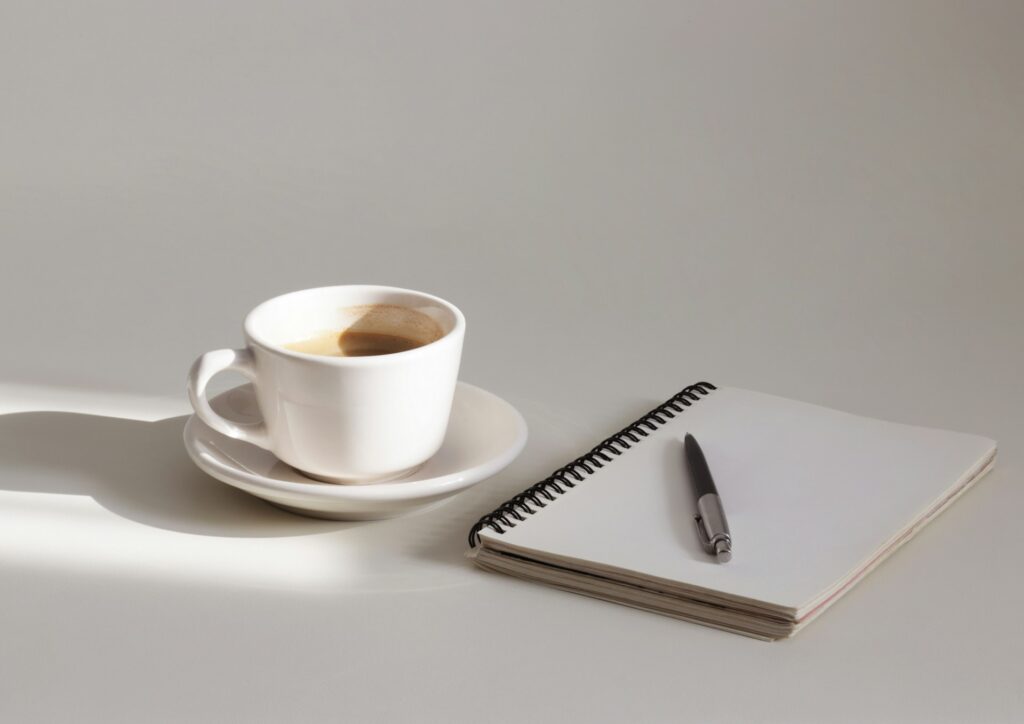 A white cup of coffee on a saucer next to a spiral notebook and a pen, cast in soft morning light on a plain background, symbolizing a calm and productive start to the day.