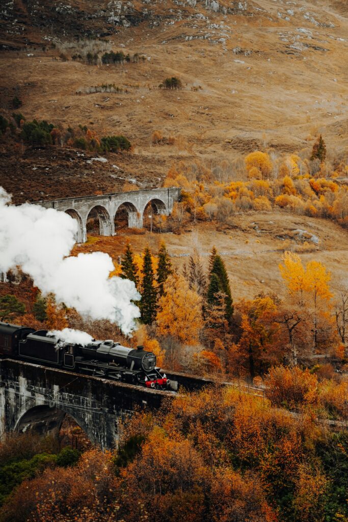 A steam train crossing an arched stone bridge surrounded by golden fall trees and rugged hills, creating a picturesque autumn landscape.