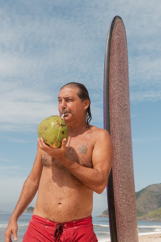 Middle-aged surfer enjoying a fresh coconut on the beach next to his surfboard