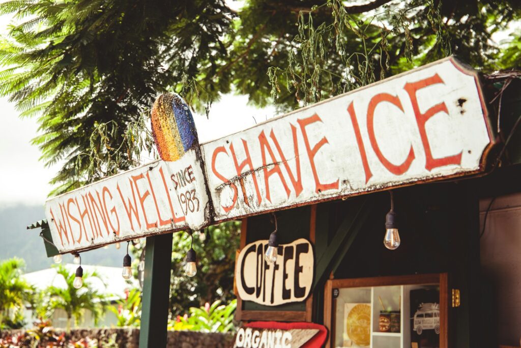 Vintage sign for Wishing Well Shave Ice and Coffee, adorned with a colorful painted surfboard and set against a tropical backdrop.