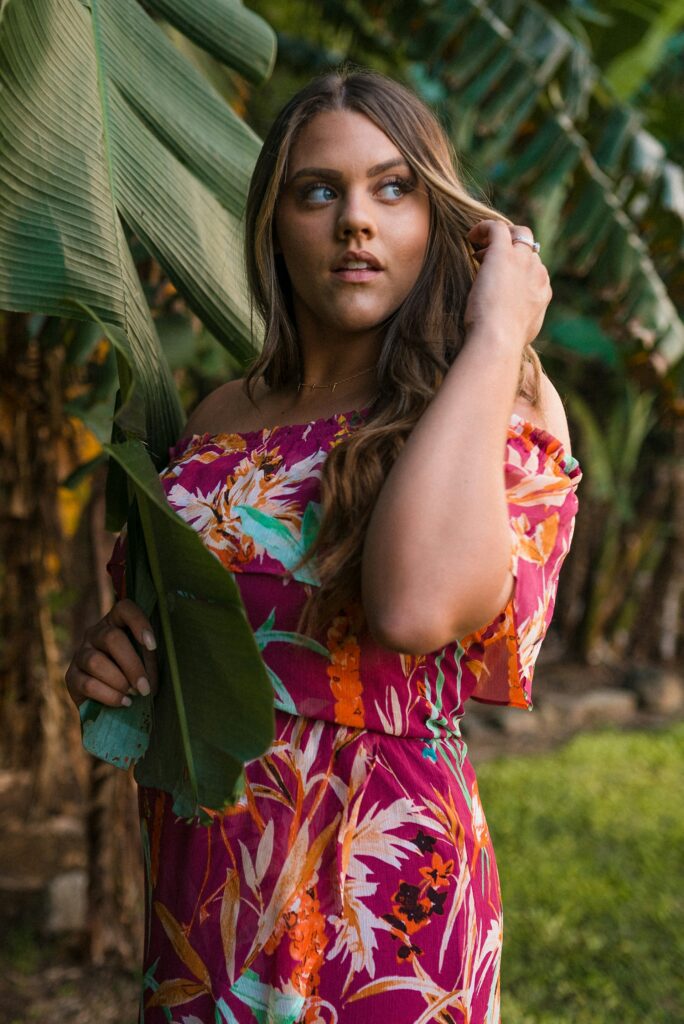 Young woman in a colorful floral dress posing in a lush Hawaiian forest, holding a green leaf.