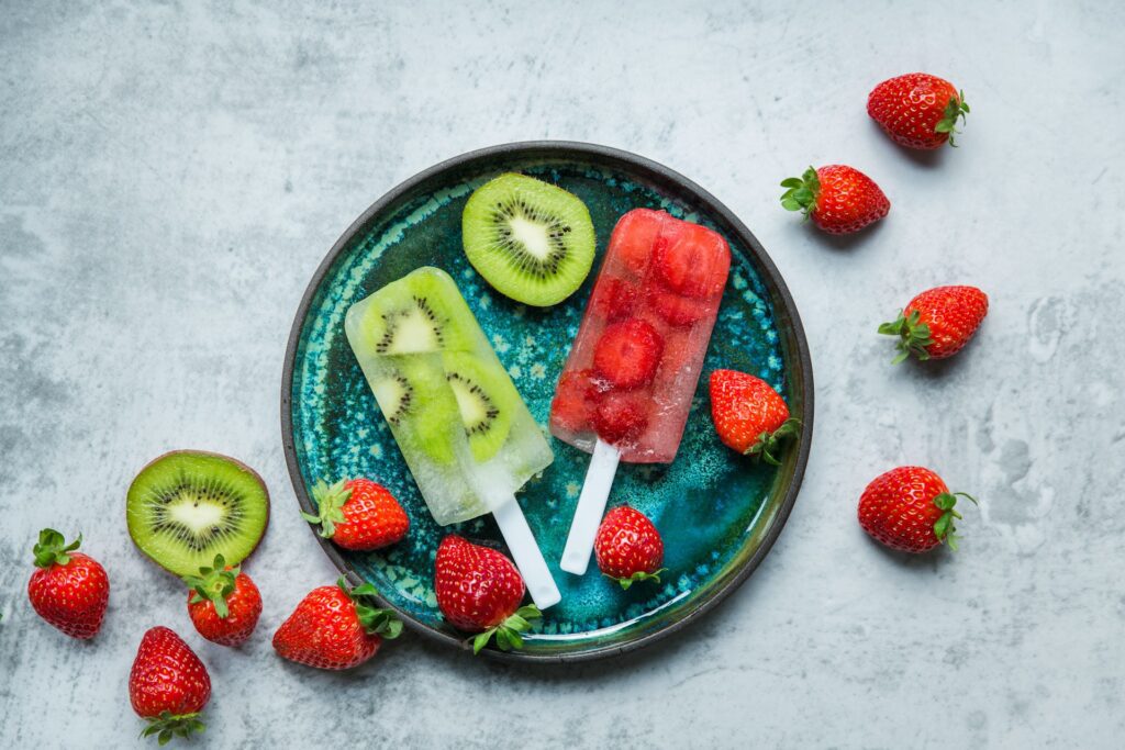 A plate of homemade frozen fruit popsicles with kiwi and strawberry flavors surrounded by fresh strawberries and kiwi slices on a textured grey background.