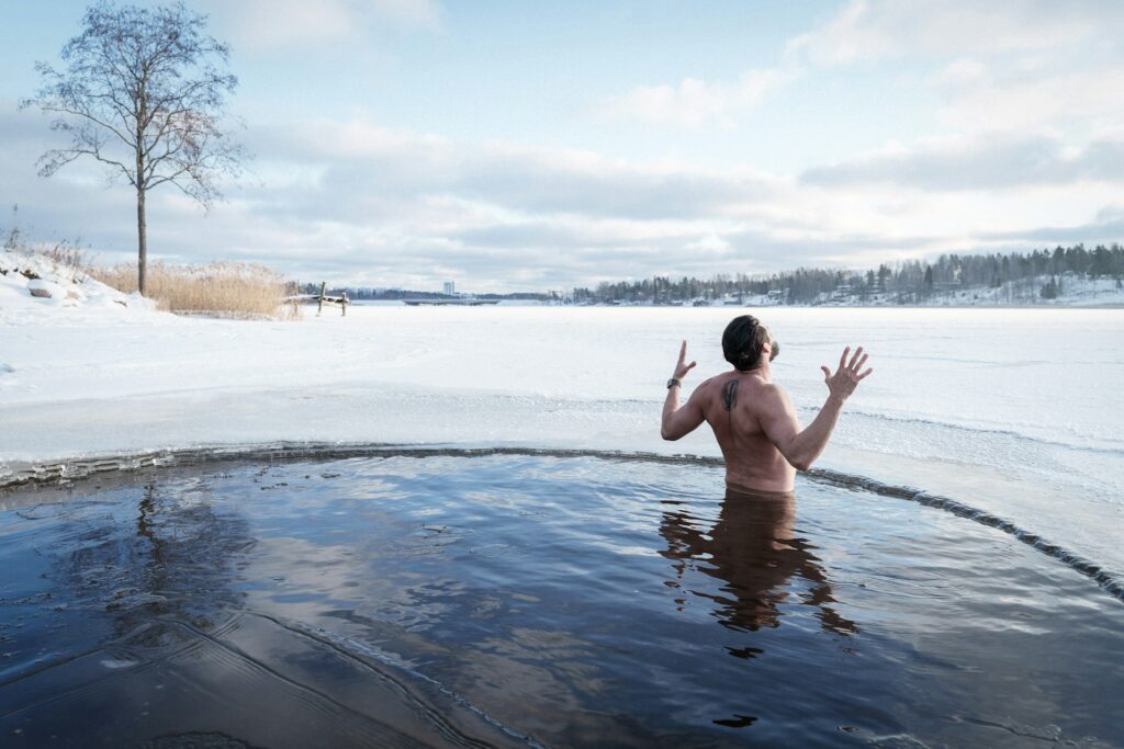 A man joyfully emerges with his hands raised from a circular hole cut in a frozen lake, surrounded by a snowy landscape.