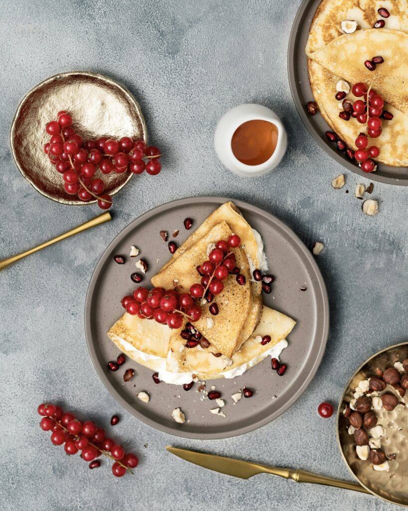 A stylishly plated breakfast of crepe-style pancakes topped with red currants, crushed nuts, and cream on a grey plate.