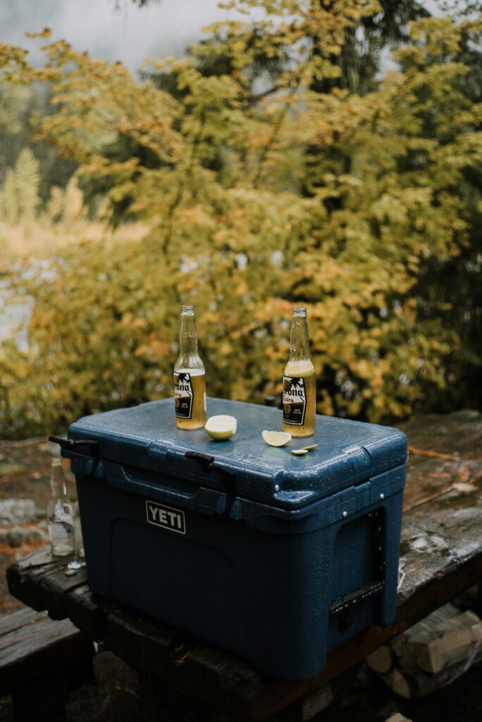 Two bottles of Corona Extra on a blue YETI cooler with slices of lime, set against a forest backdrop with autumn foliage.