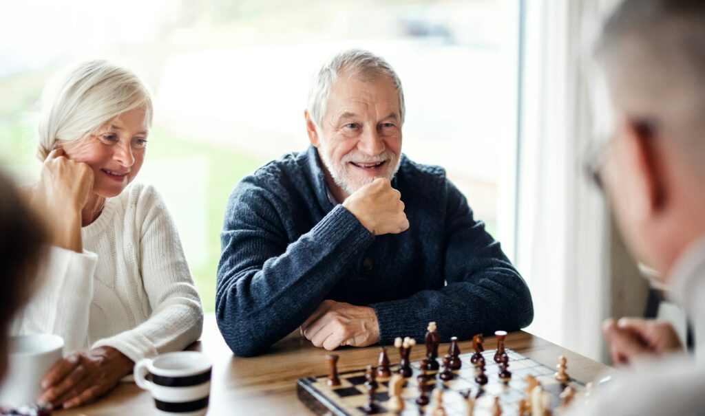 Elderly man with a beard smiling while playing chess, seated beside a woman in a white sweater, in a cozy home setting.