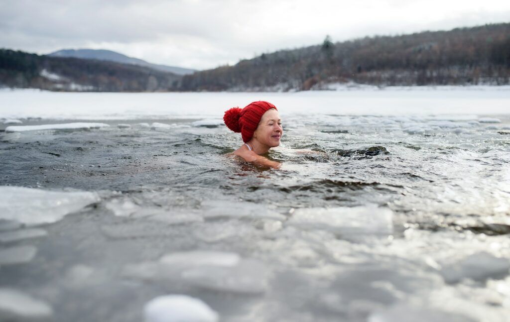 An older woman enjoys a winter swim in a natural icy water setting, smiling as she embraces the cold water challenge.