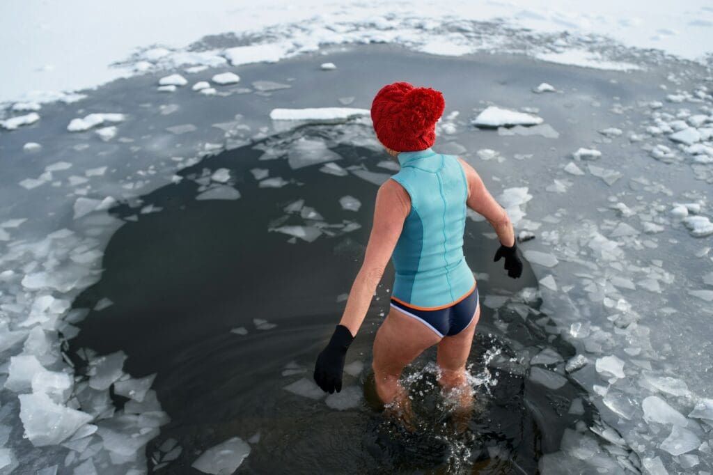 A woman in a red knit cap confidently enters an icy water hole in winter, demonstrating resilience and cold exposure therapy.
