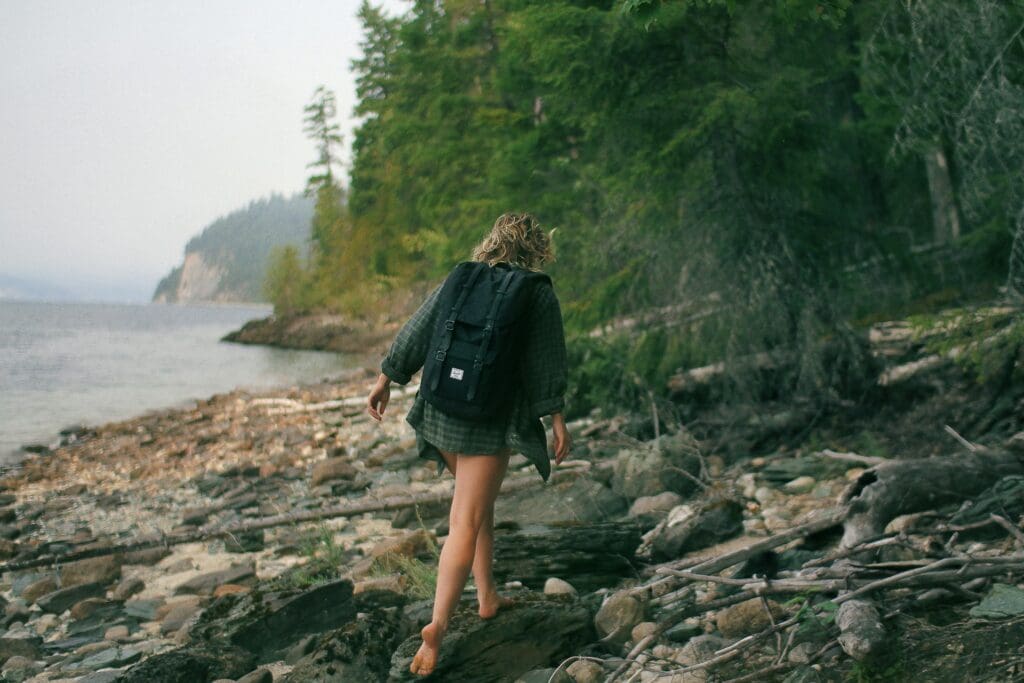 A barefoot hiker with a backpack walking along a rocky lakeside trail, surrounded by trees and misty weather.