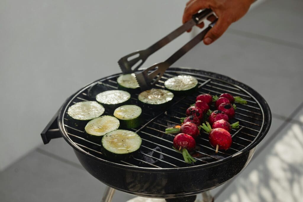 A person grilling sliced zucchini and beet kebabs on a small charcoal grill, using tongs to manage the cooking process.