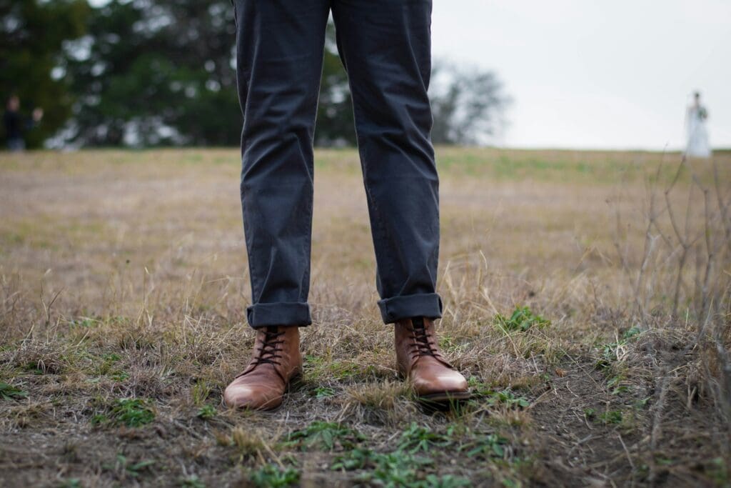 Close-up of a man's feet wearing brown leather boots, standing in a dry grass field with another person visible in the distance.