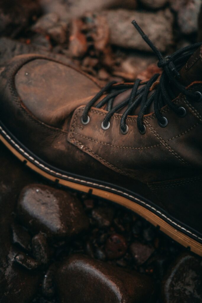 Detailed view of worn leather boots with laces, set against a background of wet, rocky ground, highlighting the boots' rugged use.