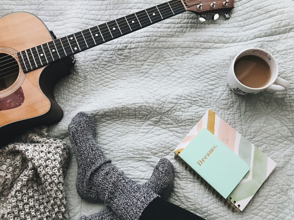 A cozy setting with a guitar lying on a quilted bed next to a person wearing wool socks, a scarf, and a notebook titled 'Dreams' with a cup of coffee.