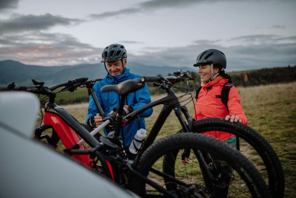 Two people, a man and a woman, preparing their mountain bikes next to their travel trailer bike rack in a field with a sunset over mountains in the background.
