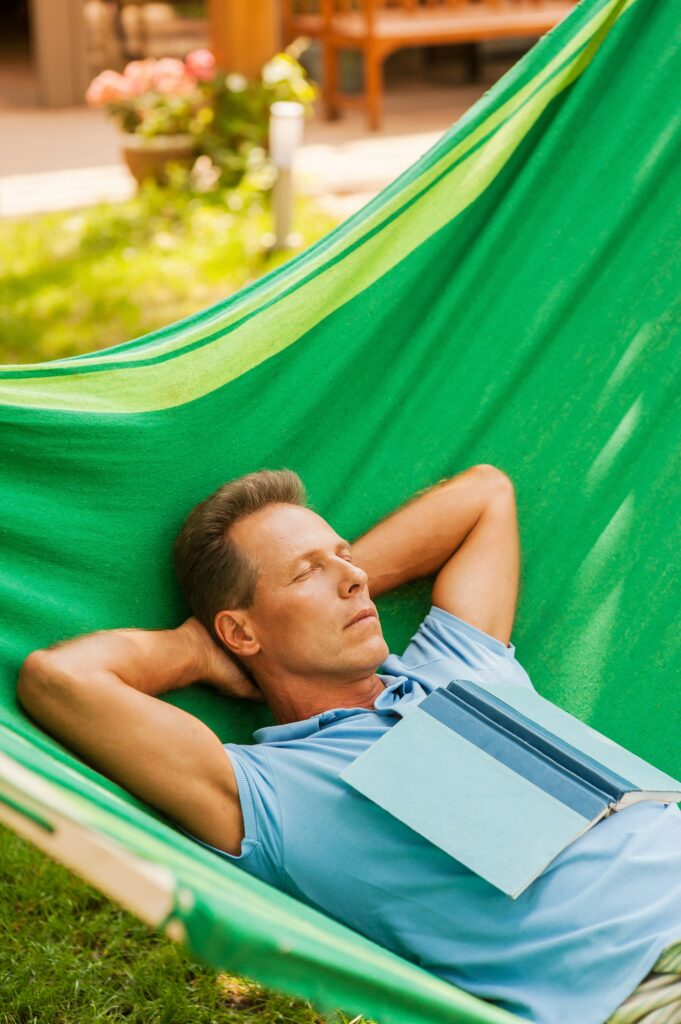 A man relaxing in a green hammock with a book on his chest, enjoying a peaceful garden setting.