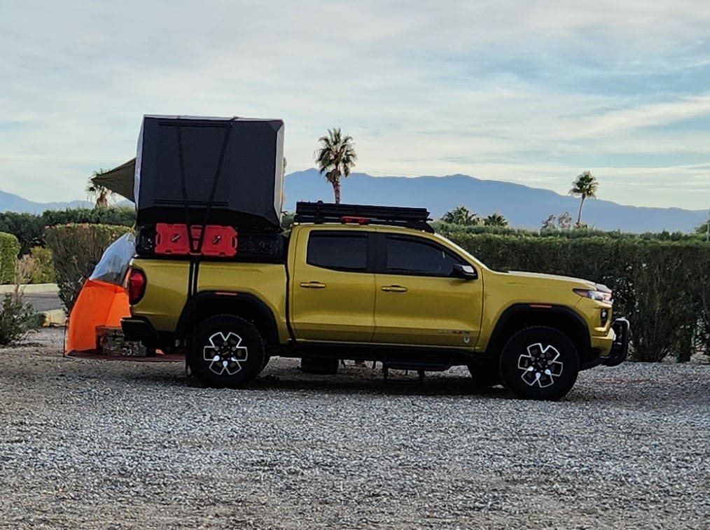 A yellow pickup truck equipped with a rooftop tent and extra storage boxes, parked in a desert setting with mountains in the background.