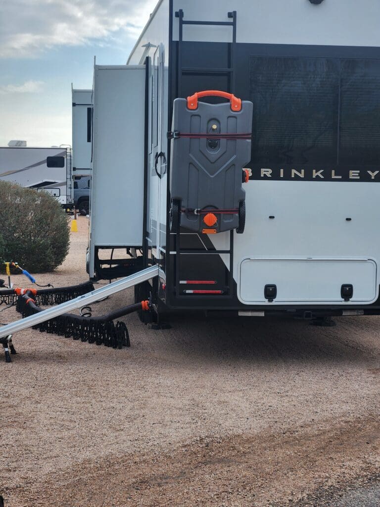 Camco portable waste tank for RV motorhomes attached to the rear ladder. RV sewer hoses are visible alongside the RV.