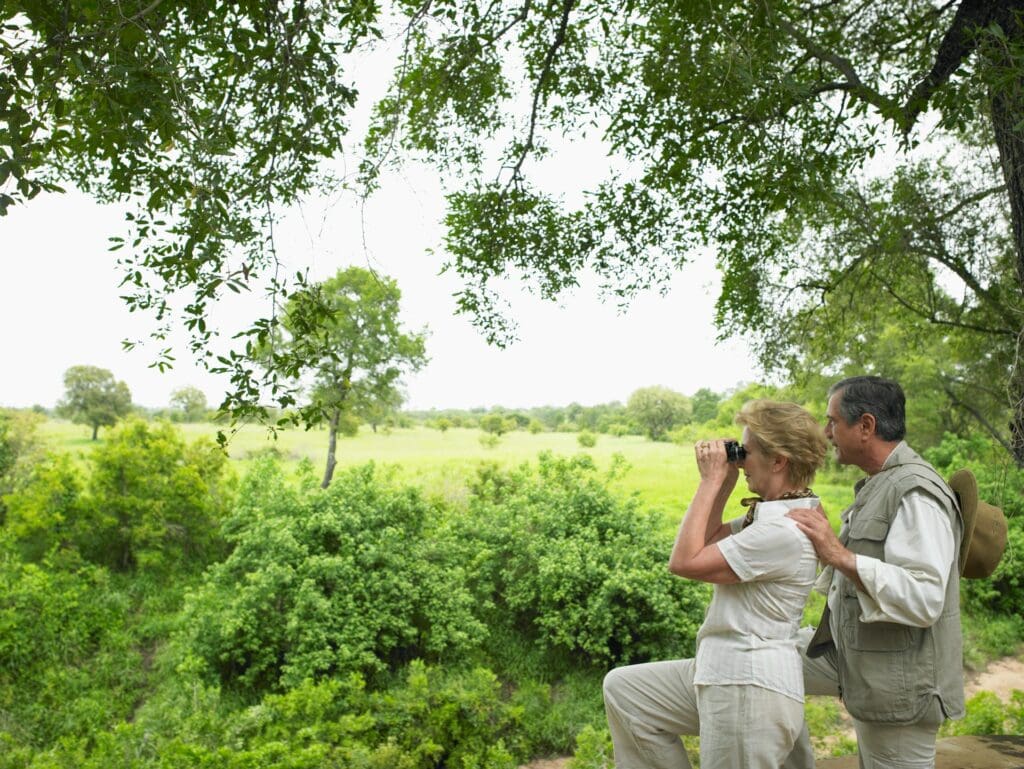 A senior couple in safari outfits using binoculars to observe wildlife from a lush, green vantage point under a tree canopy.