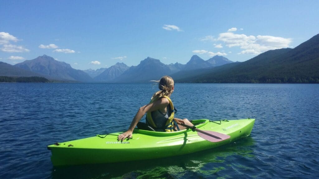 A woman in a green kayak paddles across a serene mountain lake with a backdrop of majestic mountains under a clear blue sky.