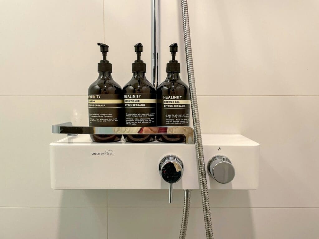 Modern bathroom shower niche featuring three dark amber bottles labeled Healinity: shampoo, conditioner, and shower gel, all in Citrus Bergamia scent, positioned on a white shower shelf with a stainless steel rail and a silver shower hose on the right.