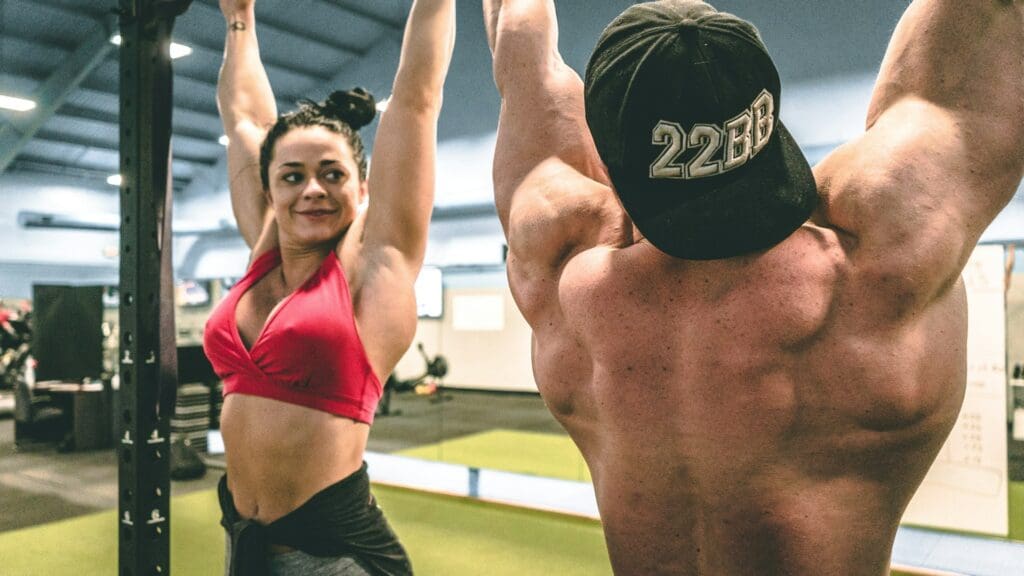Woman in a red sports bra and muscular man in a black cap doing dead hangs on pull-up bars in a gym.