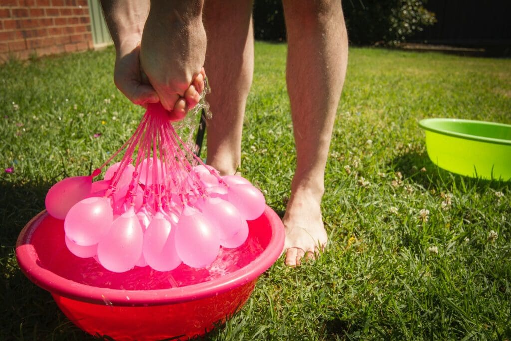 Hands gripping a cluster of pink water balloons attached to a filling hose, as water flows into a red basin on a sunny lawn.