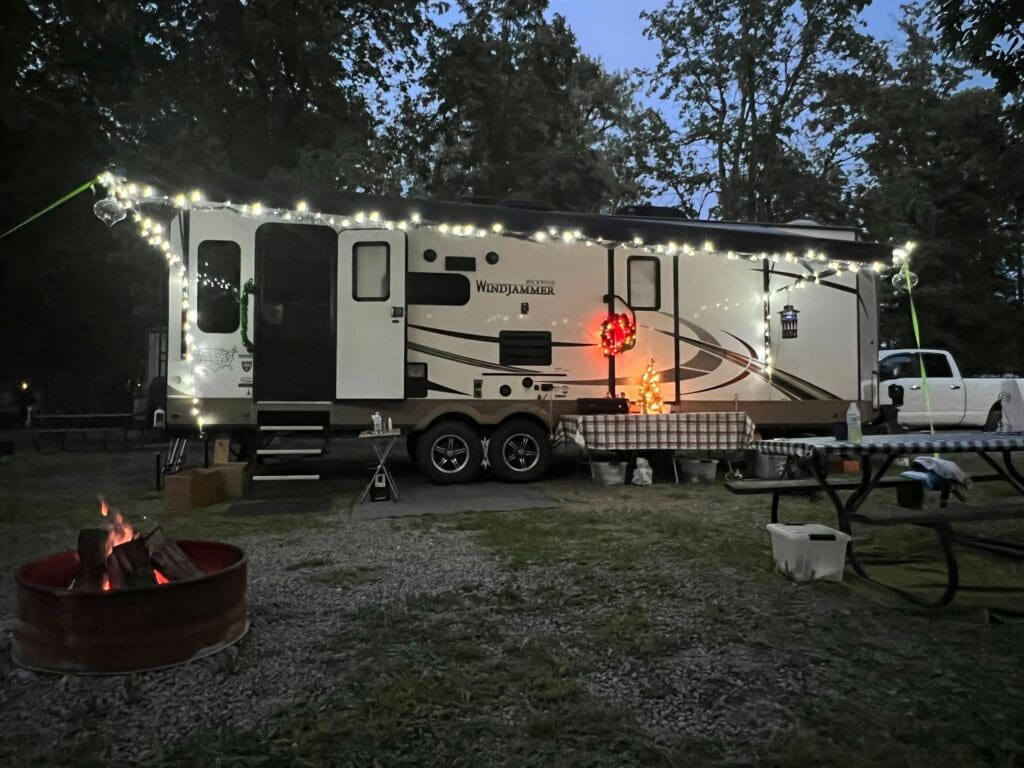 An RV travel travel at a forested campground in the evening lit up with festive lights